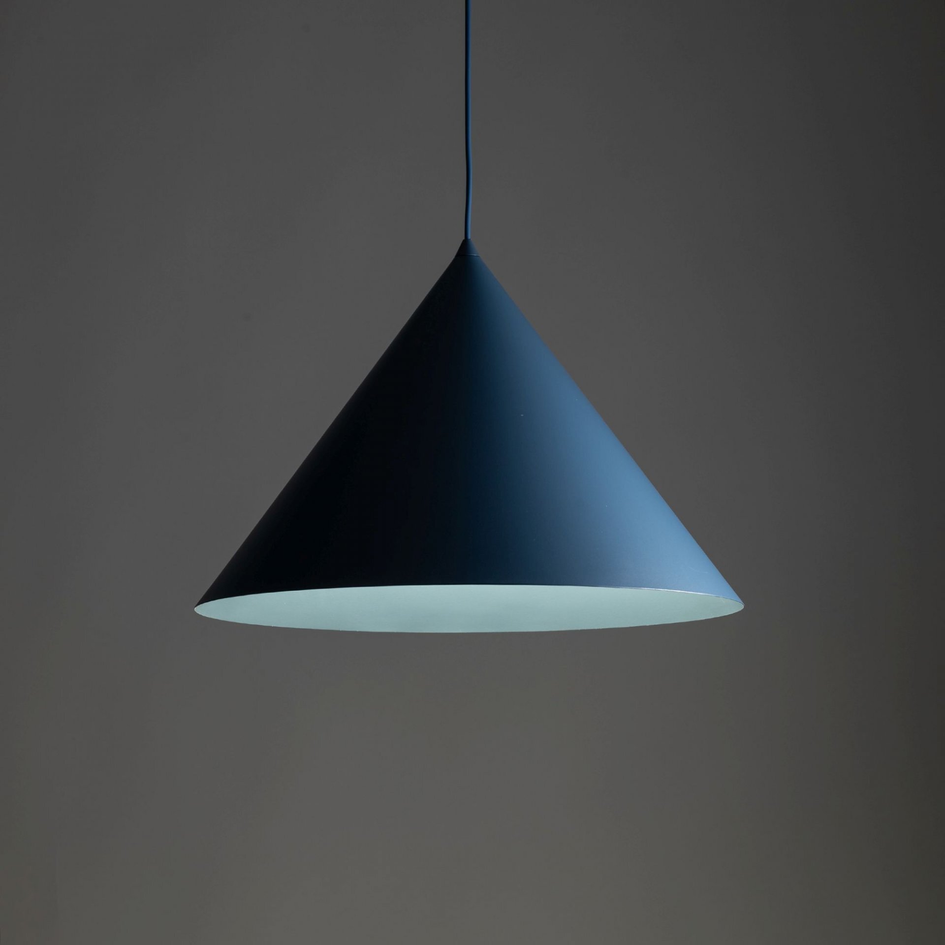 Lampa wisząca w kształcie stożka, w kolorze granatowym, zawieszona na cienkim przewodzie. Wnętrze lampy jest jasne, co kontrastuje z ciemnym zewnętrzem.