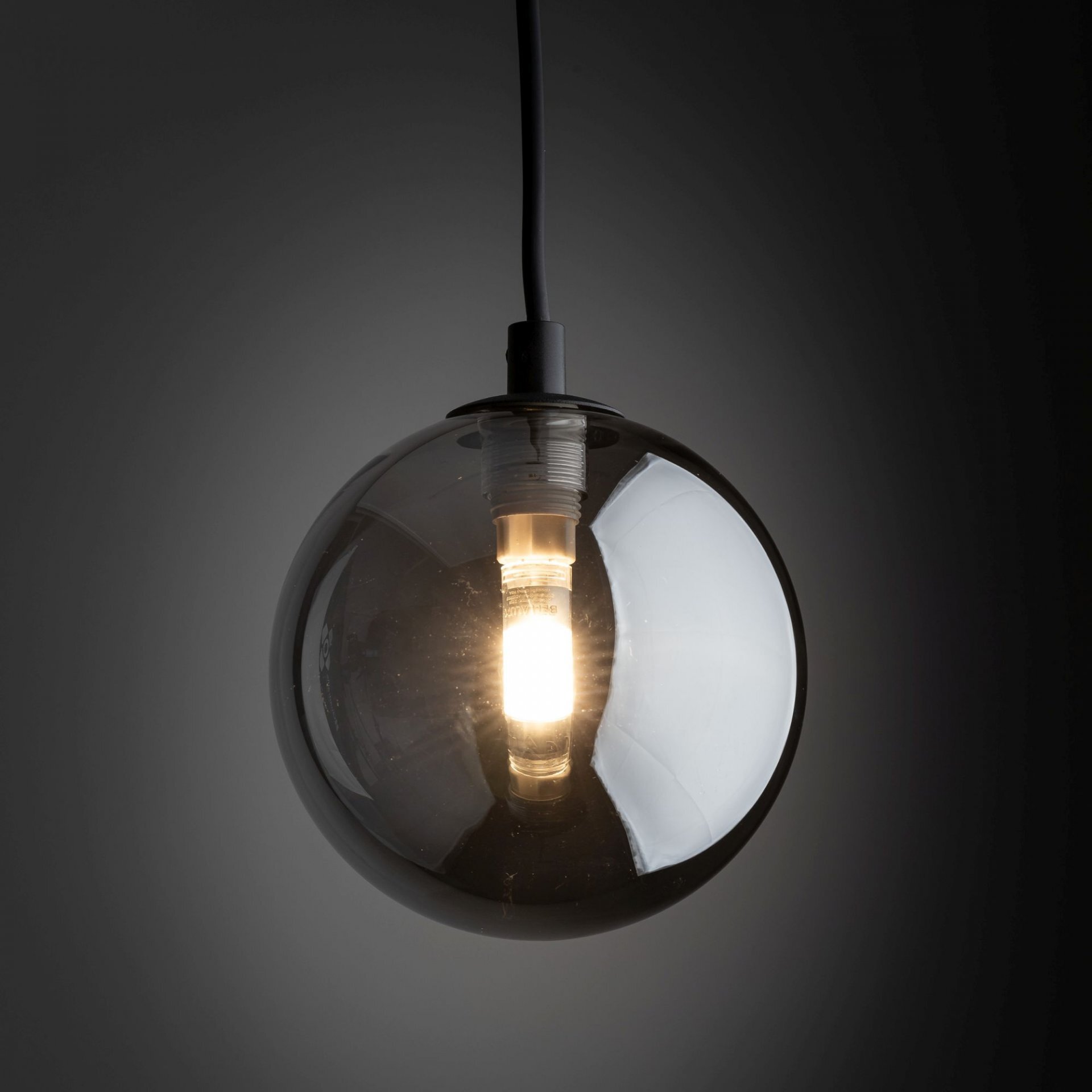 Czarna, kulista lampa wisząca z widoczną żarówką wewnątrz, emitującą ciepłe światło. Lampa zawieszona jest na czarnym przewodzie, a tło jest ciemne.
