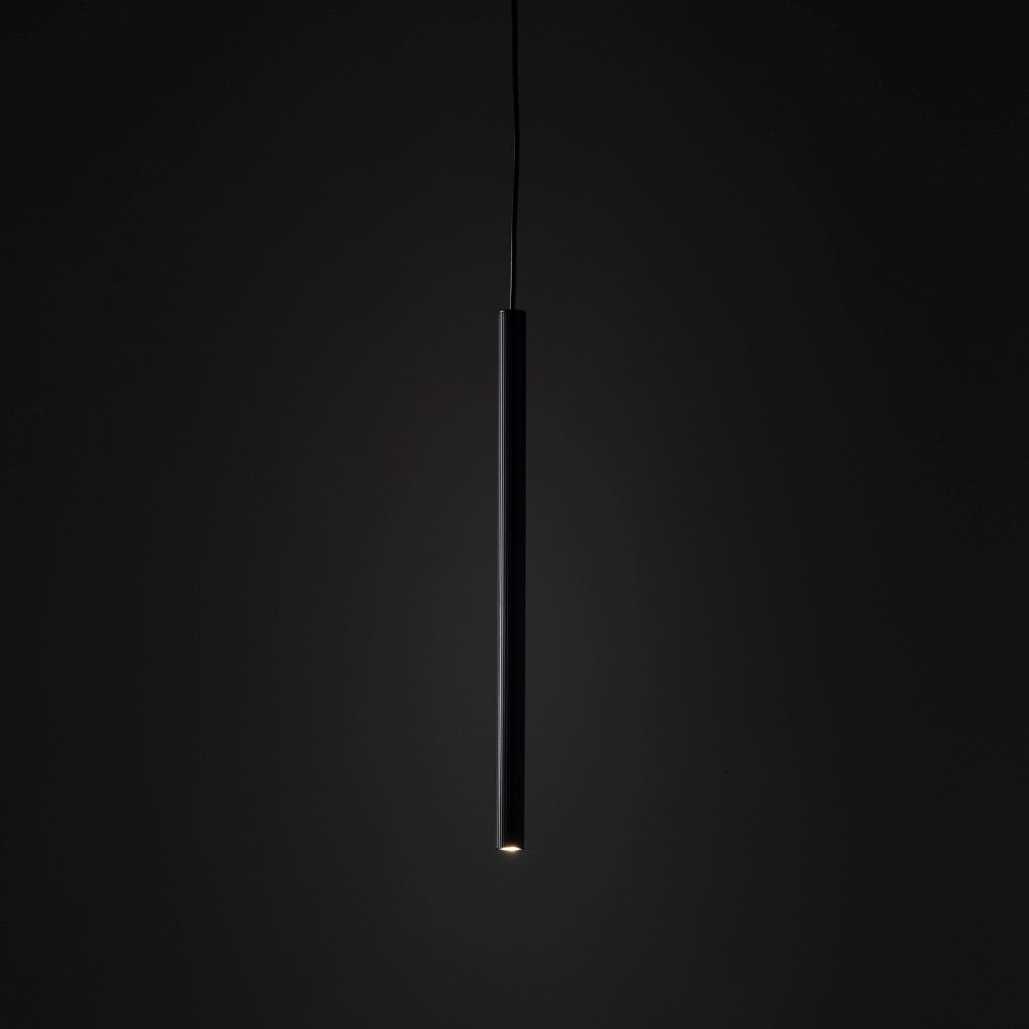 Czarna, cylindryczna lampa wisząca z pojedynczym źródłem światła na końcu, zawieszona na cienkim przewodzie. Tło jest ciemne, co podkreśla minimalistyczny design lampy.