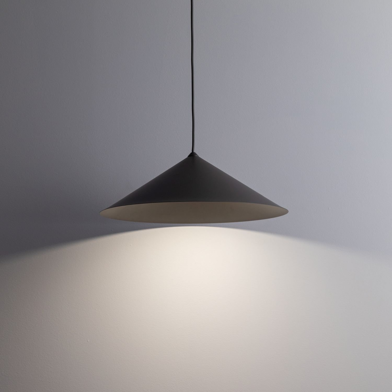 Na obrazie widoczna jest brązowa lampa wisząca o stożkowym kształcie, emitująca światło na ścianę. Lampa zawieszona jest na cienkim przewodzie.
