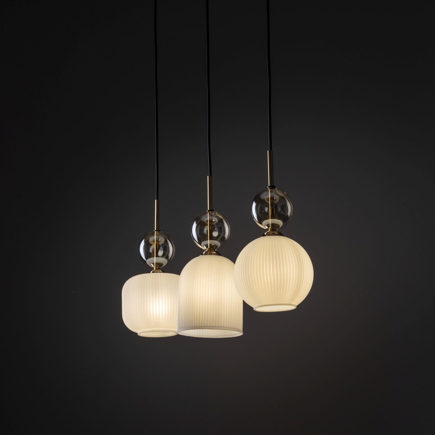 Trzy wiszące lampy z białymi kloszami o różnym kształcie, zawieszone na czarnych przewodach. Każda lampa ma złote elementy i przezroczystą kulę nad kloszem.