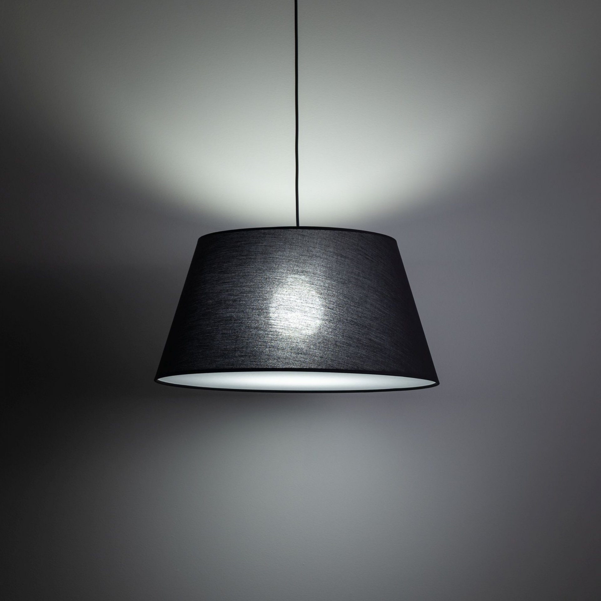 Czarna lampa wisząca z szerokim kloszem, emitująca światło na ścianę i sufit. Lampa zawieszona jest na cienkim przewodzie, a jej wnętrze jest jasne.