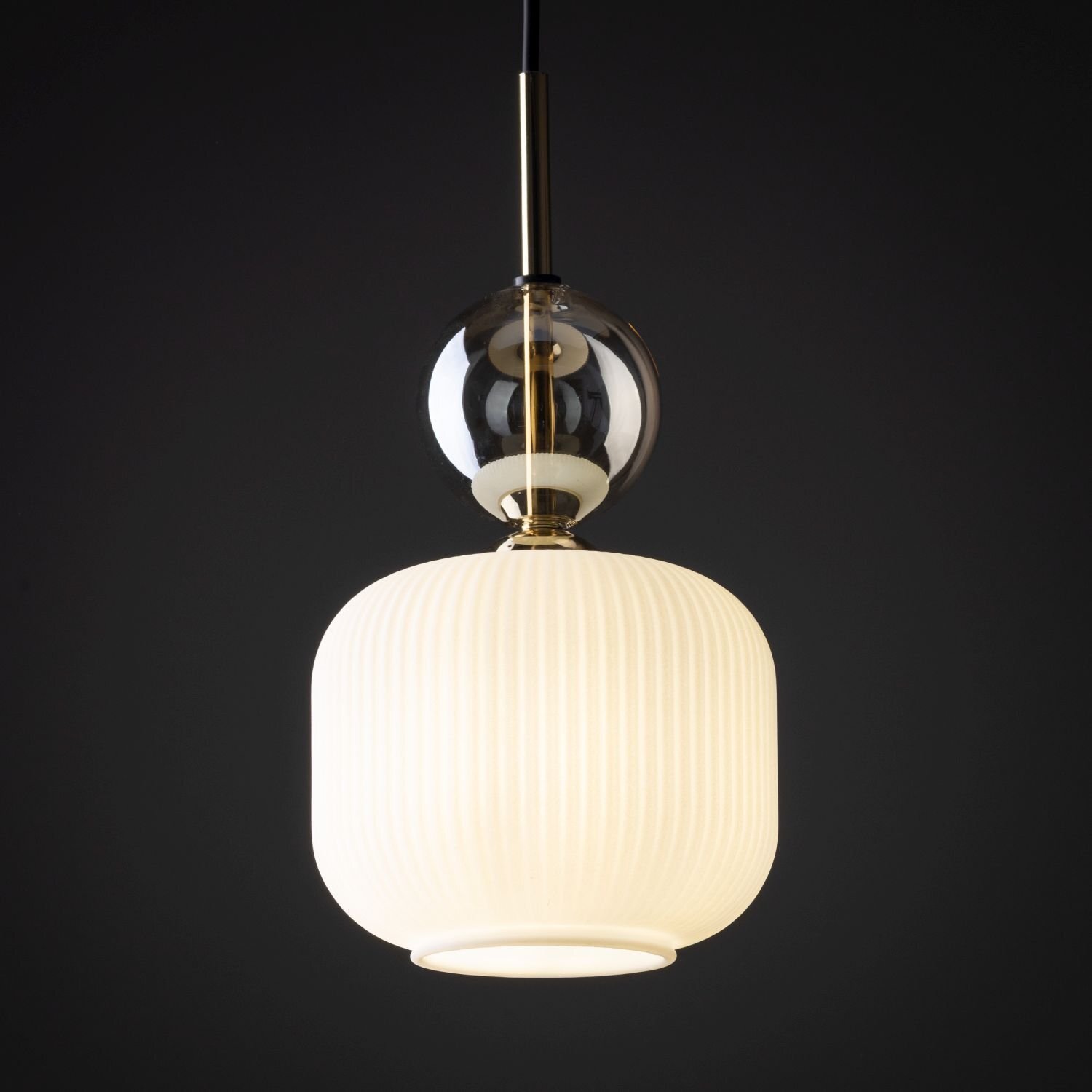 Lampa wisząca z białym, żebrowanym kloszem i złotymi elementami, zawieszona na czarnym przewodzie. Nad kloszem znajduje się przezroczysta, szklana kula.