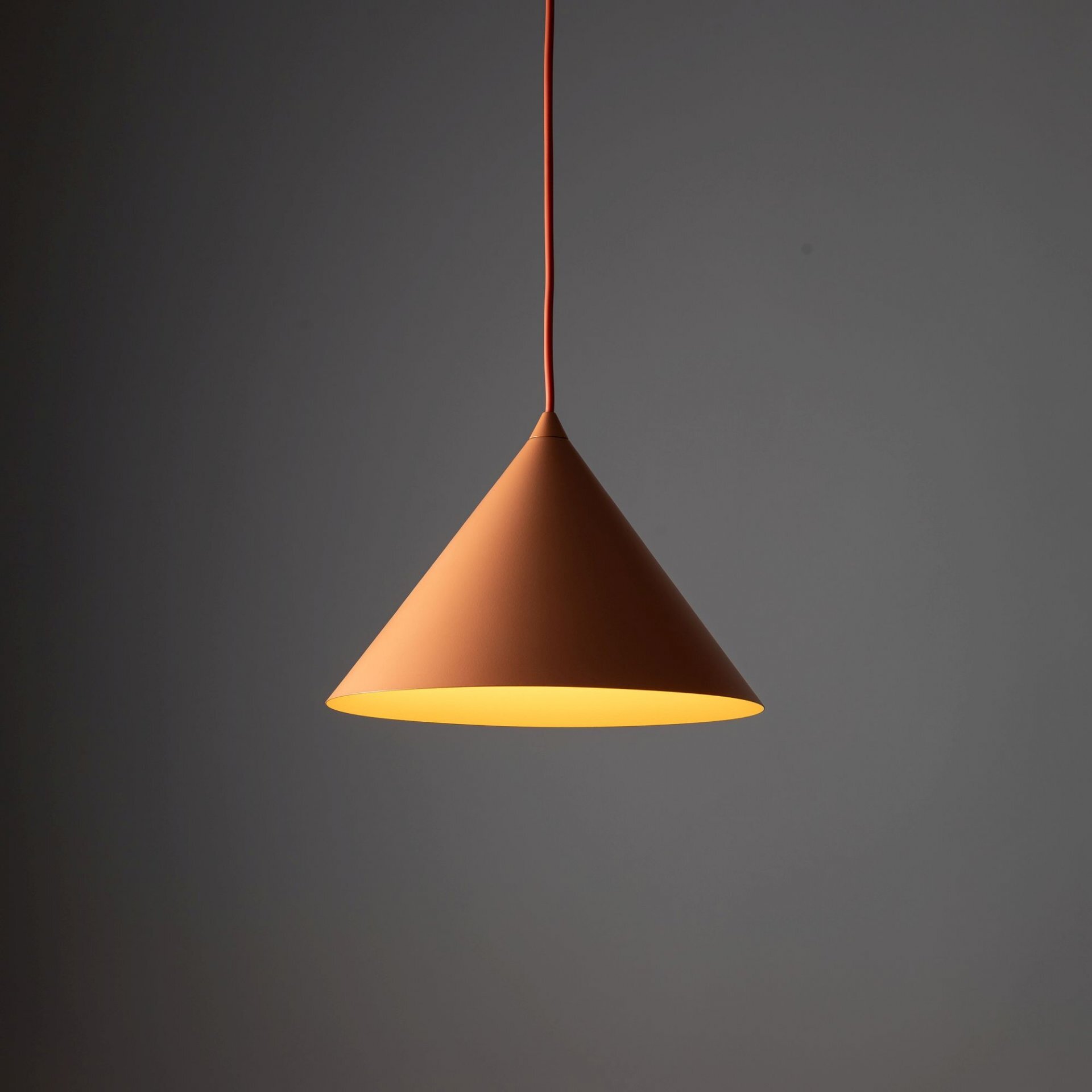 Pomarańczowa, metalowa lampa wisząca w kształcie stożka, emitująca ciepłe światło. Lampa zawieszona jest na pomarańczowym przewodzie na tle szarej ściany.