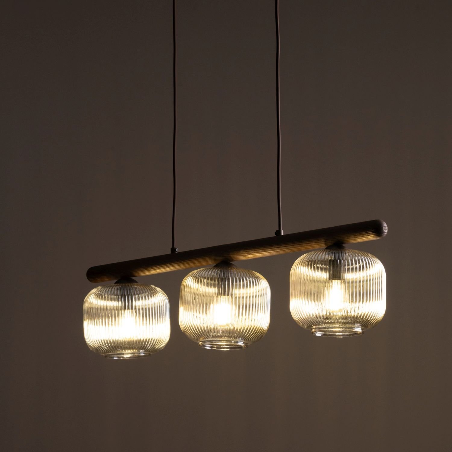 Lampa wisząca z trzema szklanymi kloszami o żebrowanej strukturze, zamontowana na poziomej belce. Klosze emitują ciepłe światło, a całość zawieszona jest na trzech cienkich przewodach.