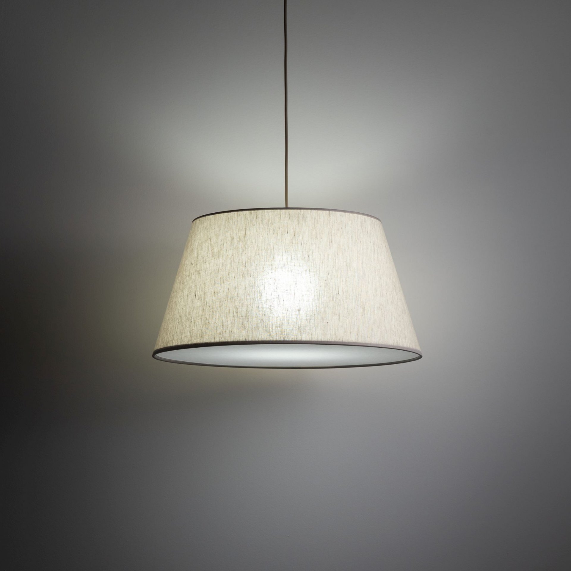 Na obrazie widoczna jest wisząca lampa z beżowym, lnianym abażurem. Lampa emituje miękkie światło, a jej kształt jest nowoczesny i prosty.