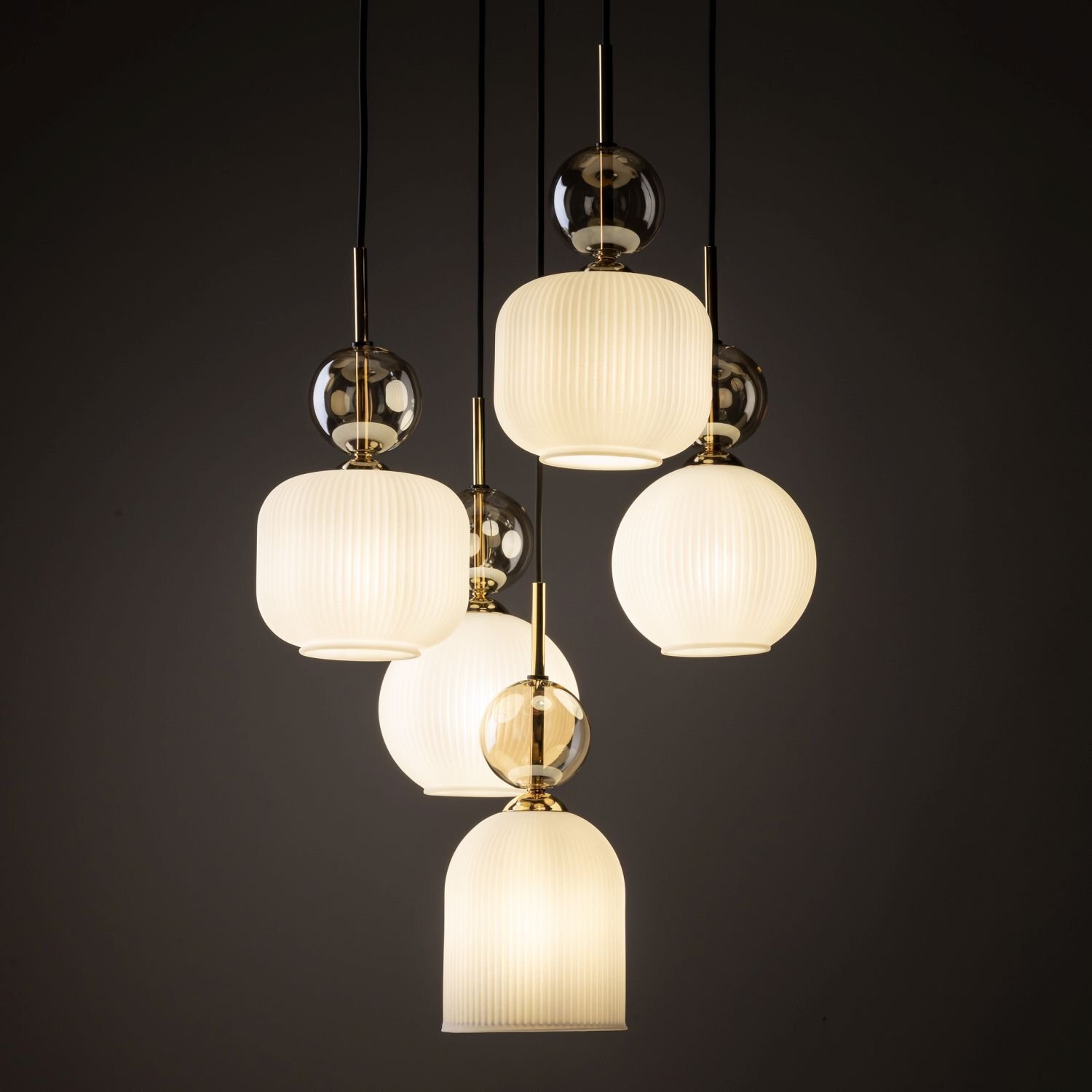 Na obrazie widoczna jest lampa wisząca z pięcioma białymi kloszami o żebrowanej strukturze, zawieszonymi na czarnych przewodach. Klosze są ozdobione złotymi elementami i przezroczystymi kulami.
