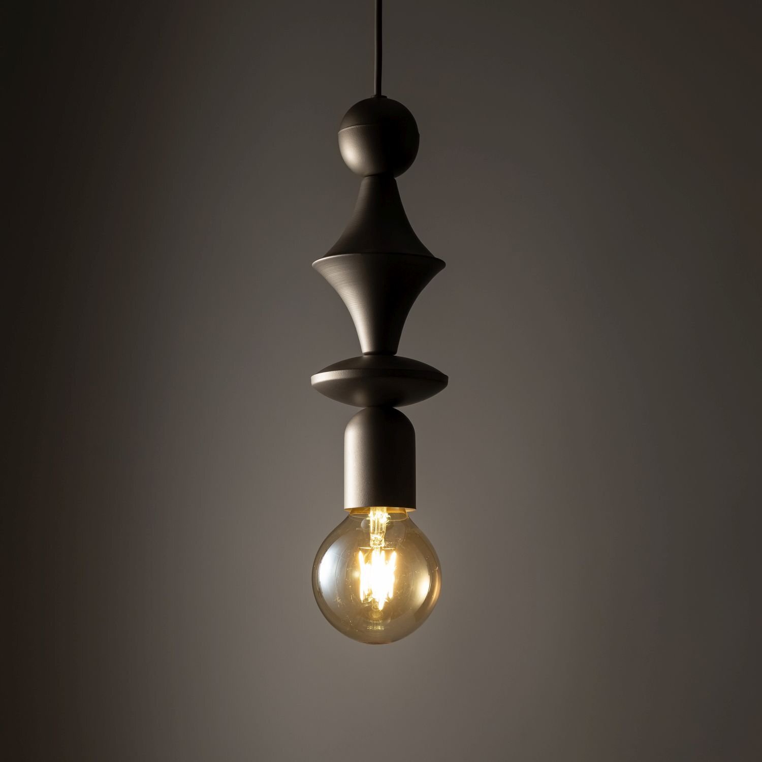 Na obrazie widoczna jest lampa wisząca z brązowej stali, z ozdobnym trzonkiem i żarówką E27. Tło jest ciemne, co podkreśla ciepłe światło emitowane przez żarówkę.