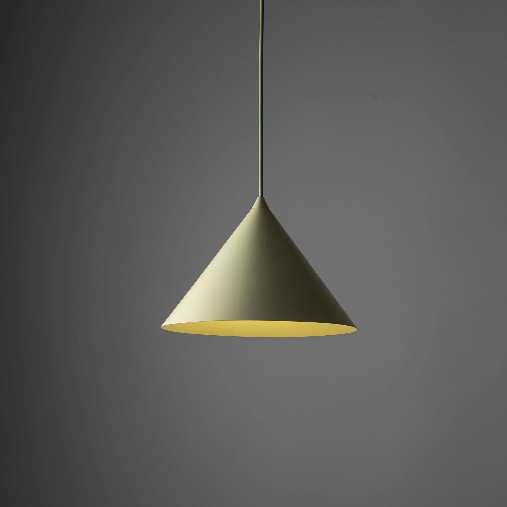 Na obrazie widoczna jest wisząca lampa w kształcie stożka, w kolorze zielonym, zawieszona na cienkim przewodzie. Tło jest jednolite, ciemnoszare.