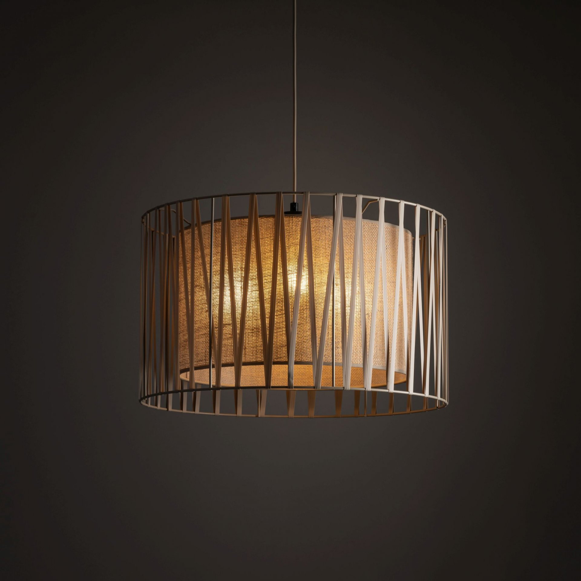 Lampa wisząca z beżowym abażurem i metalowym stelażem o geometrycznym wzorze. Światło przenika przez materiał, tworząc ciepły efekt świetlny.