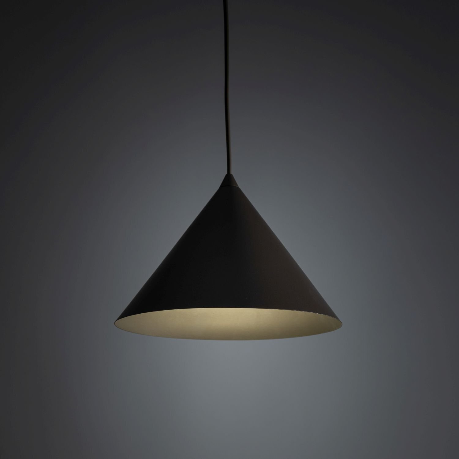 Na obrazie widoczna jest brązowa lampa wisząca o stożkowym kształcie, emitująca delikatne światło. Lampa zawieszona jest na cienkim przewodzie, a tło jest ciemne.