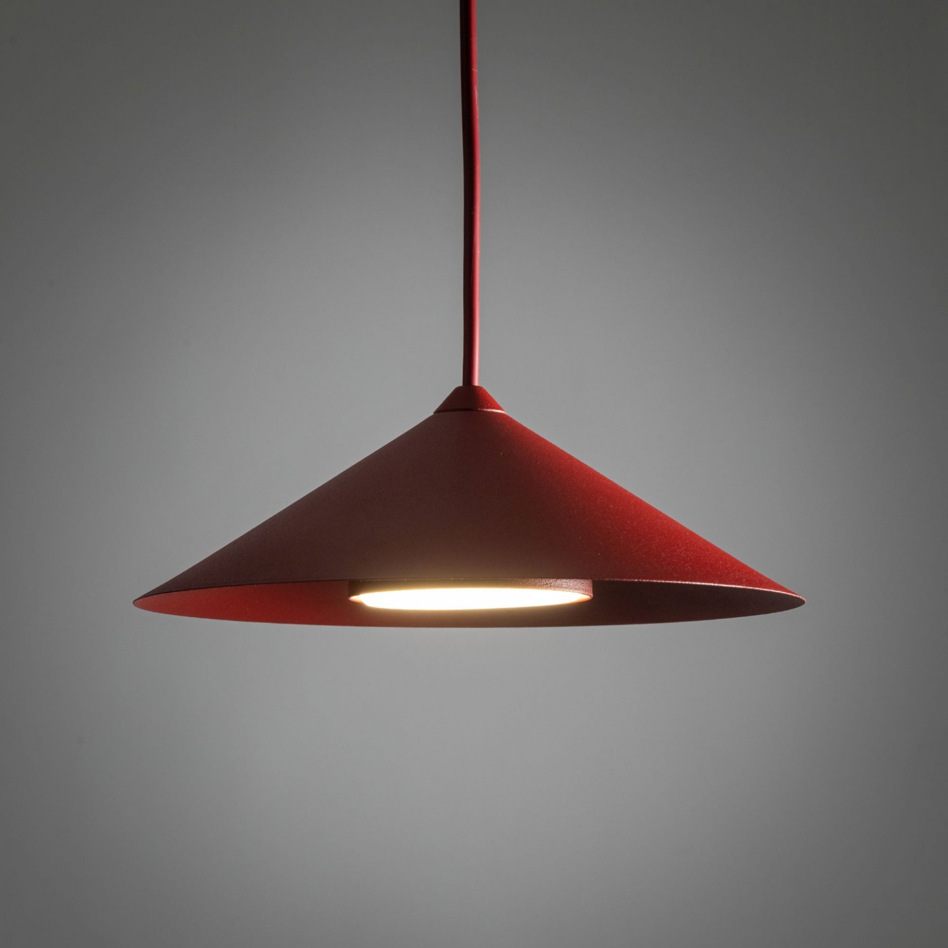 Czerwona, stożkowa lampa wisząca z widocznym źródłem światła od spodu. Lampa zawieszona jest na cienkim kablu, a tło jest szare.