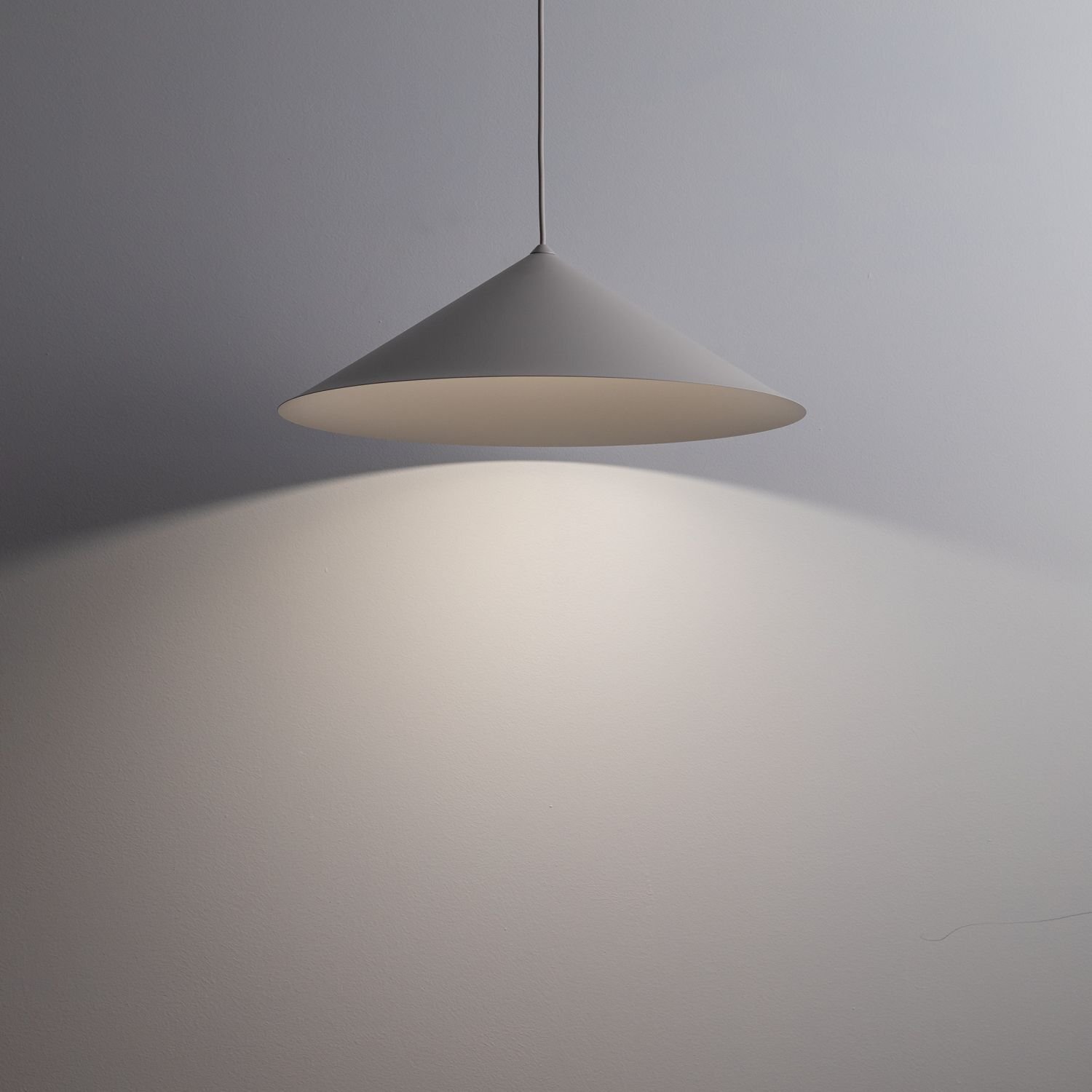 Na obrazie widoczna jest nowoczesna lampa wisząca o stożkowym kształcie, emitująca światło na ścianę. Lampa jest zawieszona na cienkim przewodzie, a jej powierzchnia ma jednolity, jasny kolor.