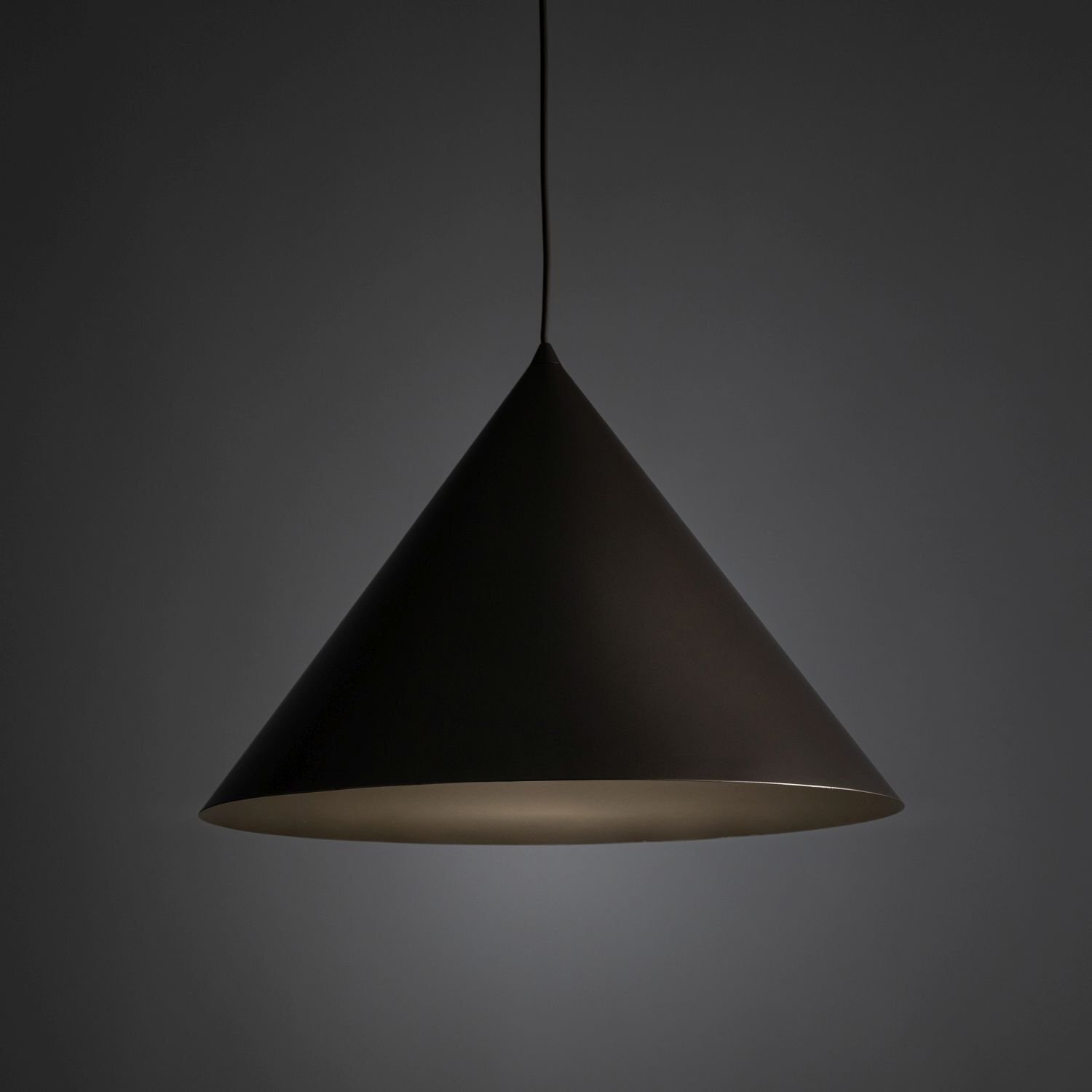 Na obrazie widoczna jest brązowa lampa wisząca w kształcie stożka, zawieszona na cienkim przewodzie. Tło jest ciemne, co podkreśla kształt i kolor lampy.