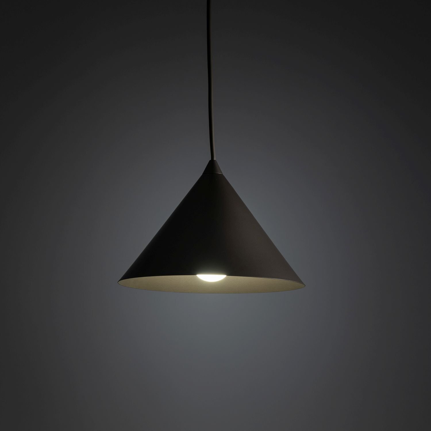 Na ciemnym tle wisi lampa o stożkowym kształcie, emitująca światło z dolnej części. Lampa jest zawieszona na cienkim przewodzie.