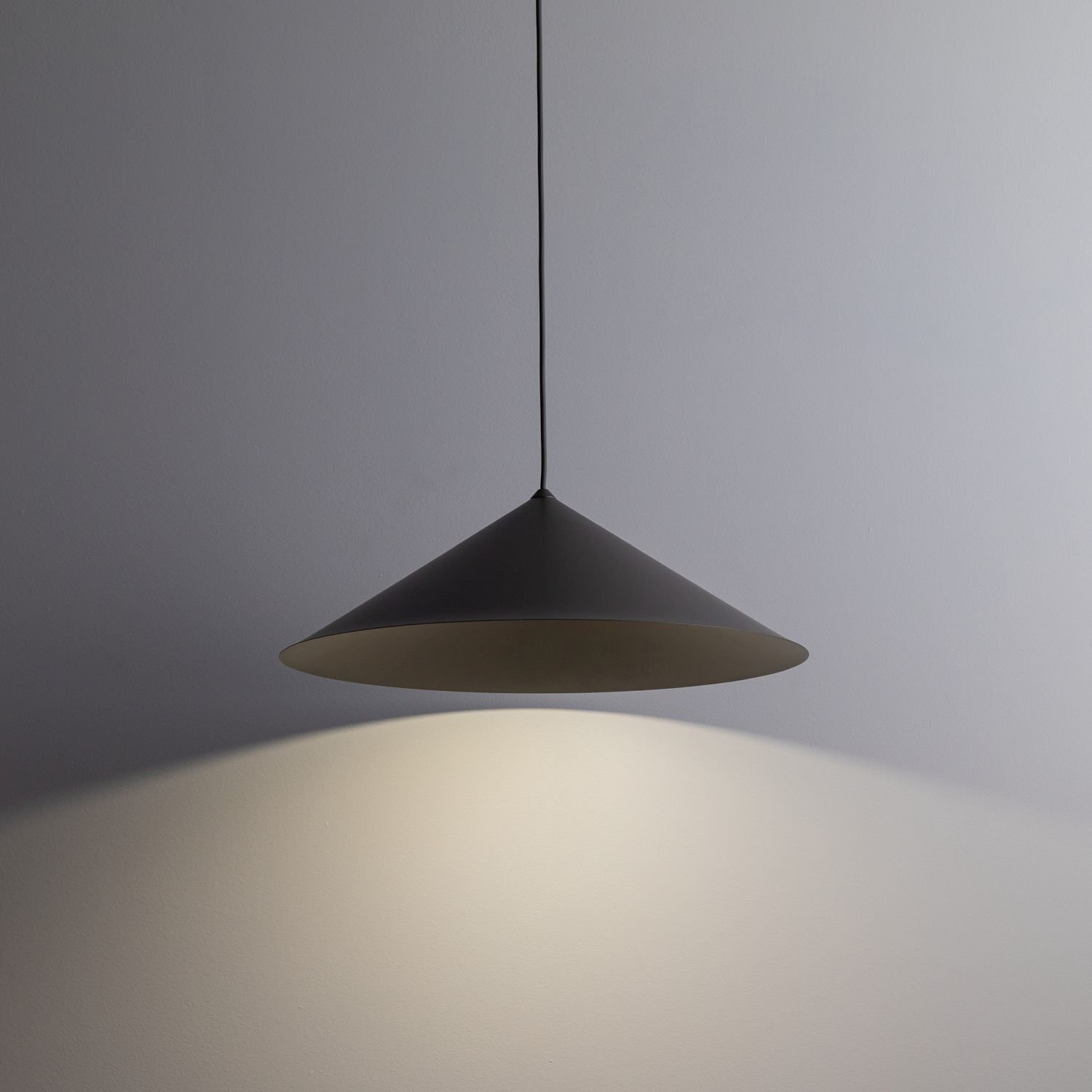 Na obrazie widoczna jest brązowa lampa wisząca o stożkowym kształcie, emitująca światło na ścianę. Lampa zawieszona jest na cienkim przewodzie.