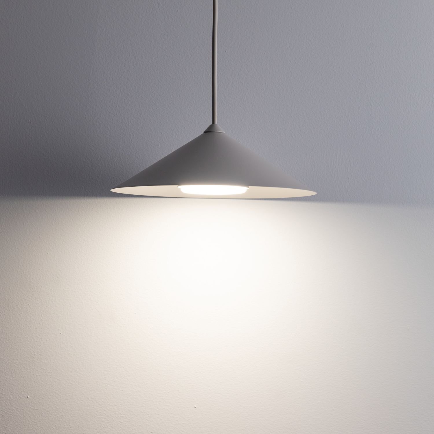 Na obrazie widoczna jest wisząca lampa o stożkowym kloszu w kolorze beżowym, emitująca jasne światło na ścianę. Lampa jest zawieszona na cienkim przewodzie.