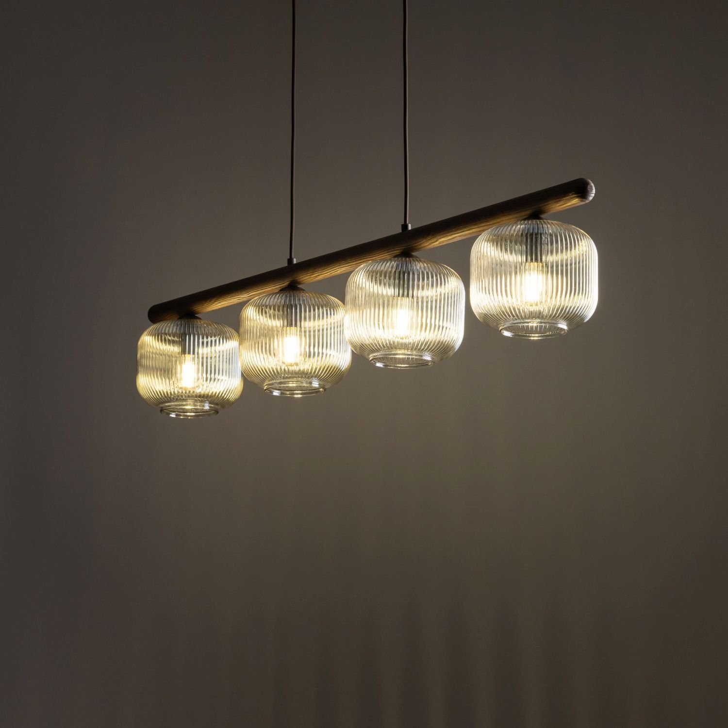 Na obrazie widoczna jest lampa wisząca z czterema szklanymi kloszami o żebrowanej strukturze, zawieszona na metalowym pręcie. Każdy klosz emituje ciepłe, żółte światło.