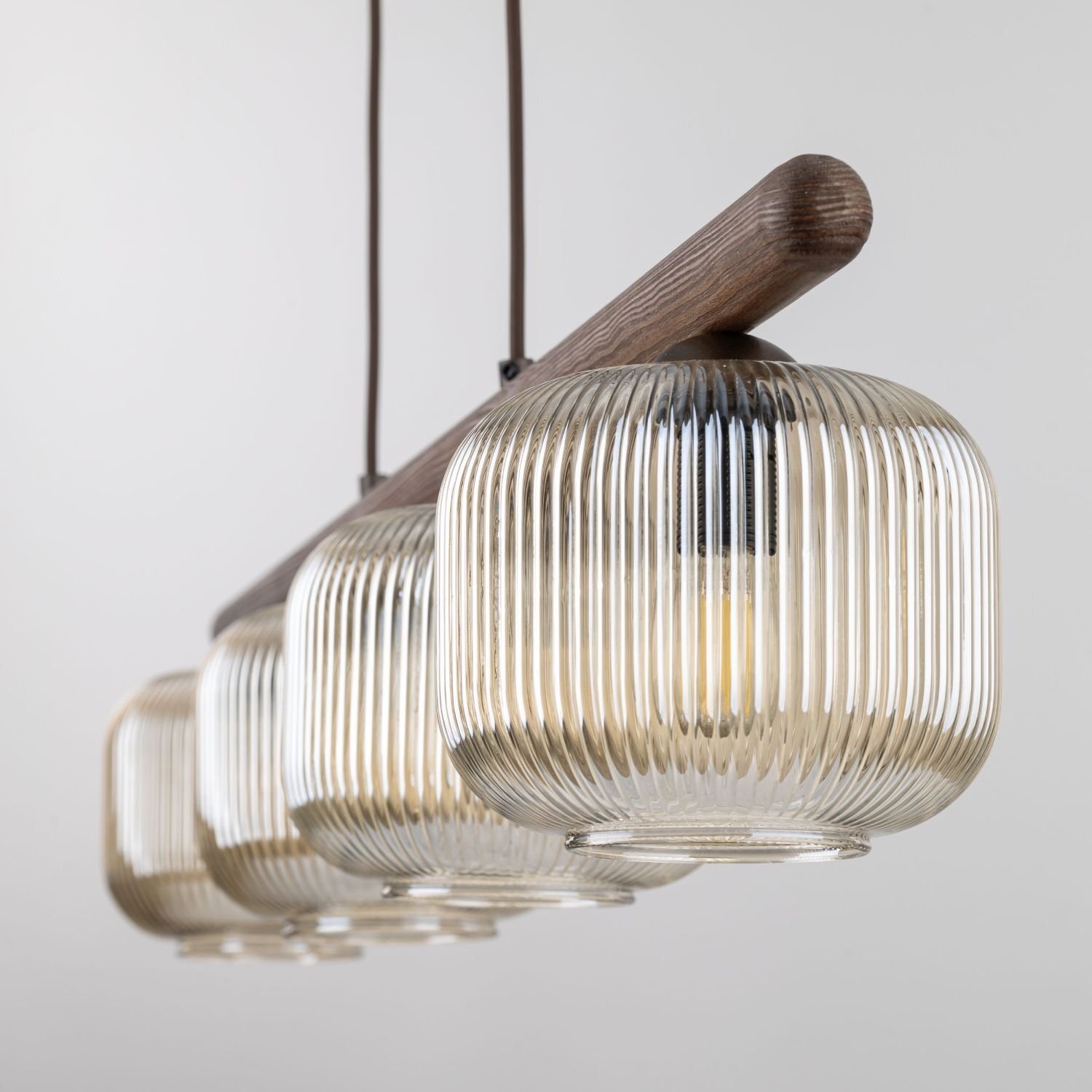 Lampa wisząca z czterema żarówkami w szklanych kloszach o żebrowanej strukturze, zamocowana na drewnianej belce. Klosze mają delikatny, brązowy odcień.