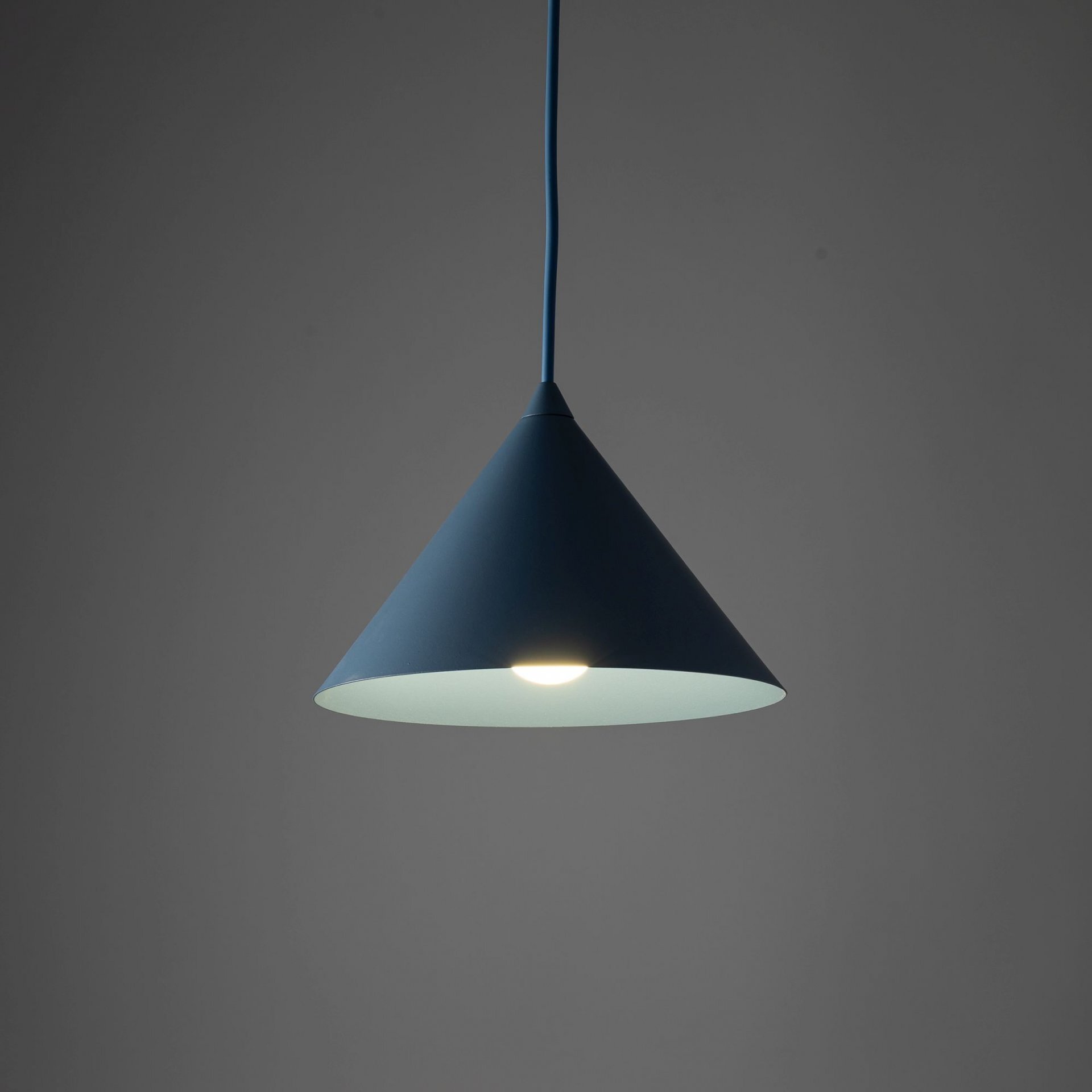 Na obrazie widoczna jest granatowa lampa wisząca o stożkowym kształcie, emitująca światło. Tło jest jednolite i ciemne.