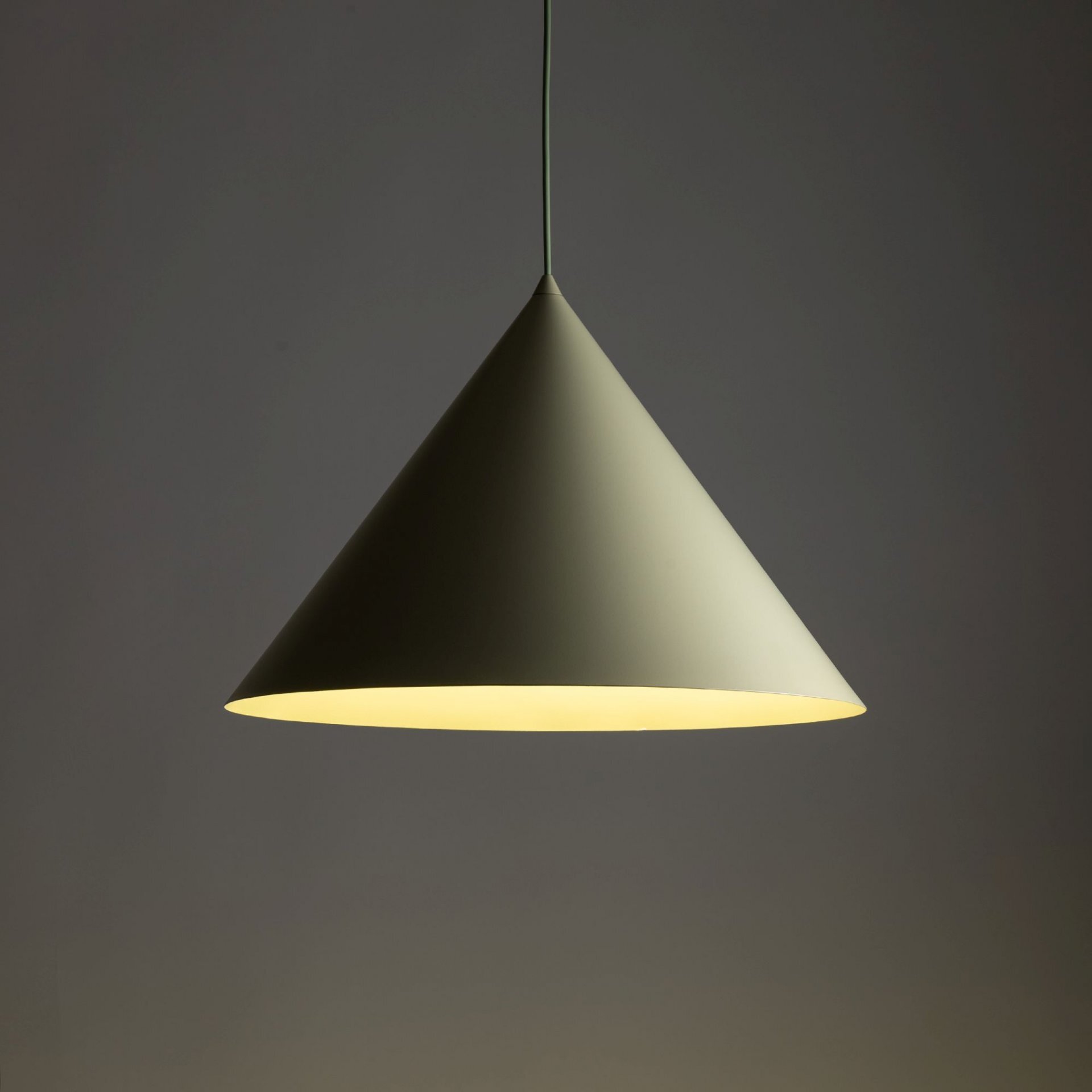 Na obrazie widoczna jest stożkowa lampa wisząca w kolorze pistacjowym, emitująca ciepłe światło. Lampa zawieszona jest na cienkim przewodzie, a tło jest jednolite i ciemne.