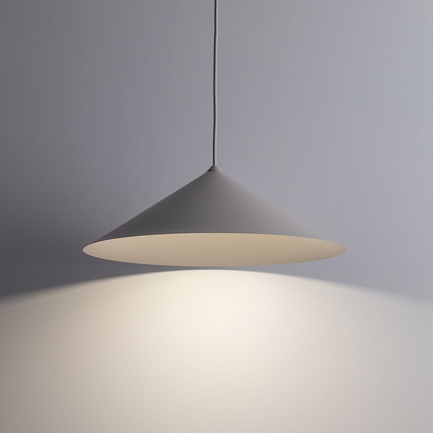Na obrazie widoczna jest nowoczesna lampa wisząca o stożkowym kształcie w kolorze kaszmiru, emitująca światło na ścianę. Lampa zawieszona jest na cienkim przewodzie, a jej powierzchnia jest gładka.