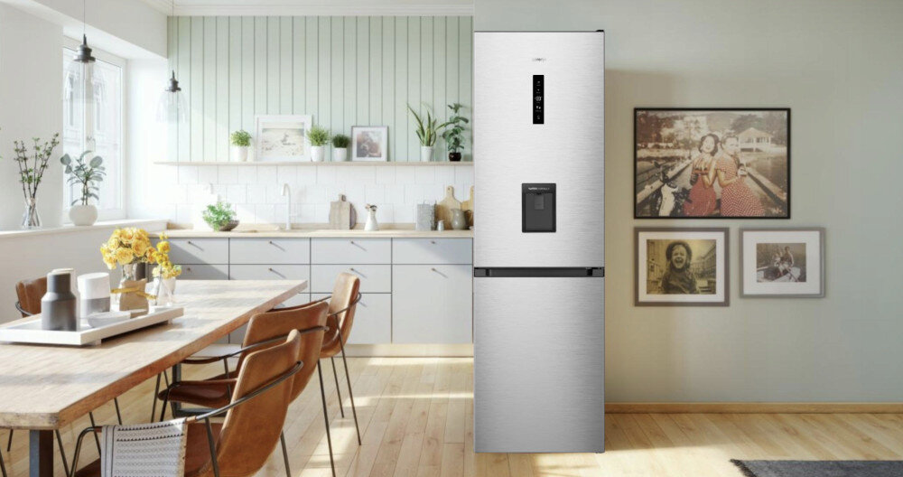 W jasnej kuchni stoi szara lodówka z wbudowanym dystrybutorem wody. Obok lodówki na ścianie wiszą trzy obrazy w ramkach.