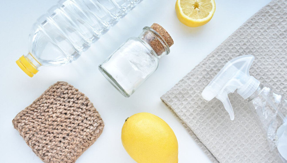 Na białym tle znajdują się plastikowa butelka z wodą, szklany słoik z korkiem, połówka cytryny, cała cytryna, przezroczysty spryskiwacz, beżowa ściereczka i brązowa, pleciona podkładka.