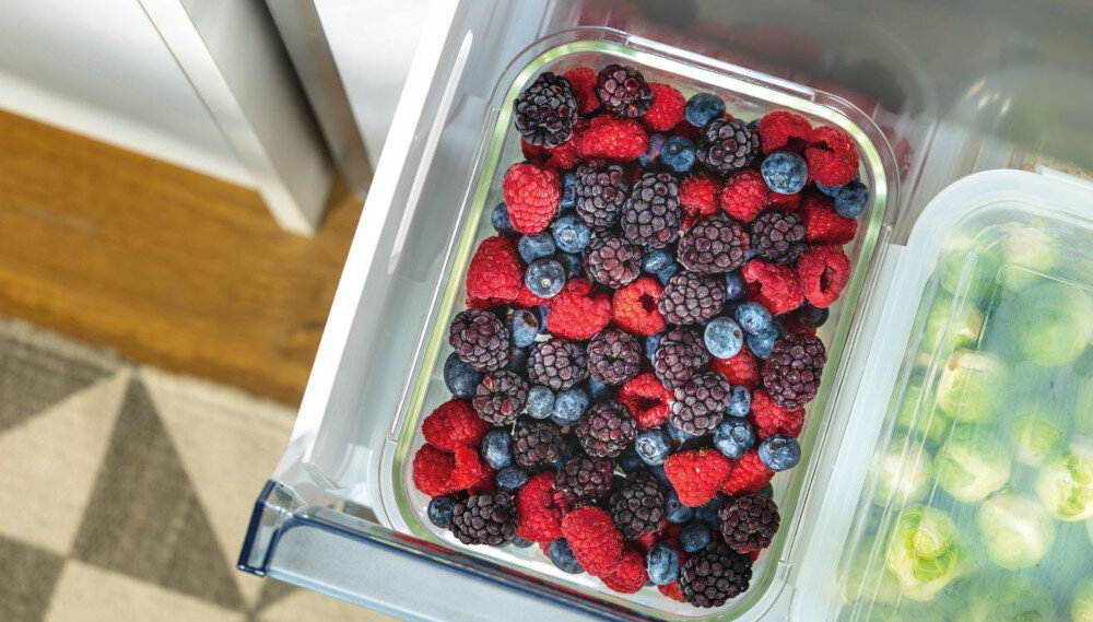W szufladzie lodówki znajduje się pojemnik z mieszanką mrożonych owoców, w tym malin, jeżyn i borówek. Obok widoczny jest fragment innego pojemnika z zielonymi warzywami.