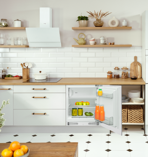 Kuchnia z białymi szafkami i drewnianym blatem, na którym stoją różne naczynia i dekoracje. Otwarta lodówka zawiera warzywa i napoje, a na półkach znajdują się słoiki z przyprawami i talerze.