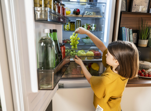 Osoba stoi przed otwartą lodówką, sięgając po kiść winogron. W lodówce widoczne są różne produkty spożywcze, w tym butelki i owoce.