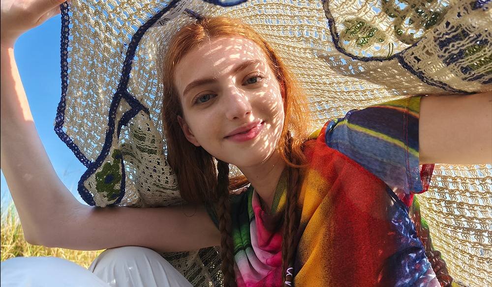 Osoba trzyma nad głową ażurową tkaninę, przez którą prześwituje światło. Ubrana jest w kolorową koszulkę, a w tle widać niebo.