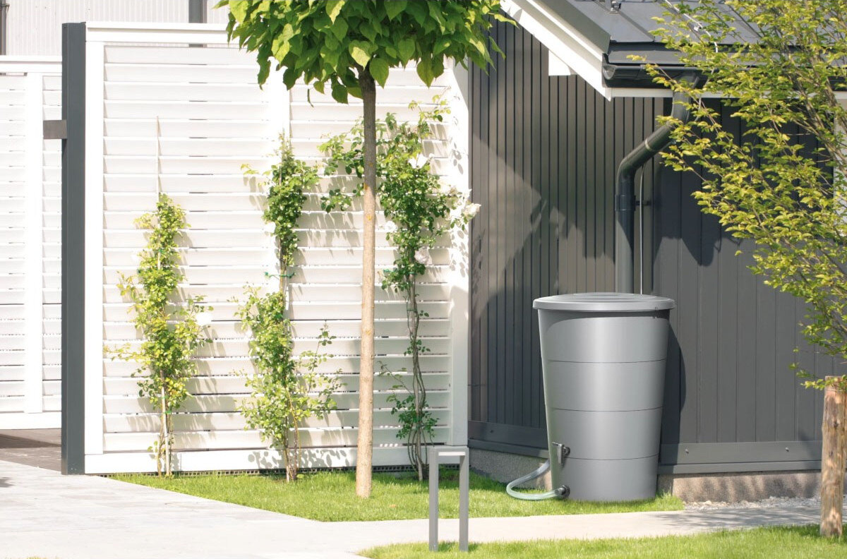 Na obrazie widoczny jest antracytowy zbiornik na deszczówkę ustawiony przy ścianie budynku, z podłączonym wężem. W tle znajduje się drewniany płot oraz rośliny ozdobne.