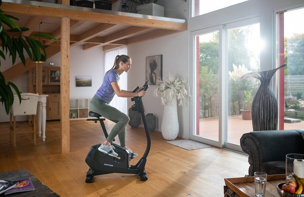 Osoba ćwiczy na rowerze magnetycznym KETTLER Ride 100 w jasnym, nowoczesnym wnętrzu z dużymi oknami. Na rowerze widoczne jest logo \'KETTLER\'.
