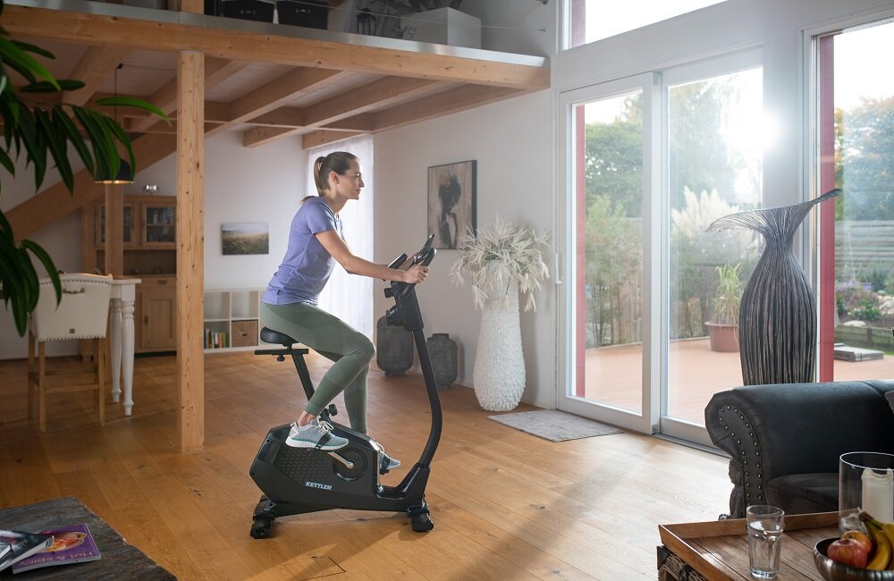 Osoba ćwiczy na rowerze magnetycznym KETTLER Ride 100 w jasnym, nowoczesnym wnętrzu z dużymi oknami. W tle widoczne są drewniane elementy wystroju i rośliny ozdobne.