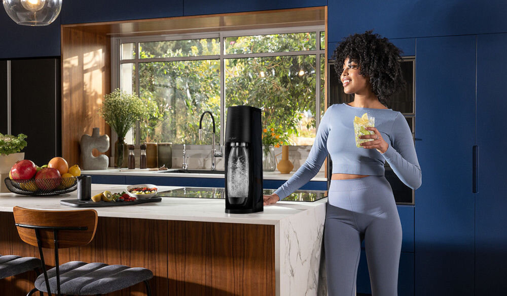 Na blacie kuchennym stoi czarny saturator z przezroczystą butelką wypełnioną wodą gazowaną. W tle widoczne są okna z widokiem na zieleń oraz miska z owocami na blacie.