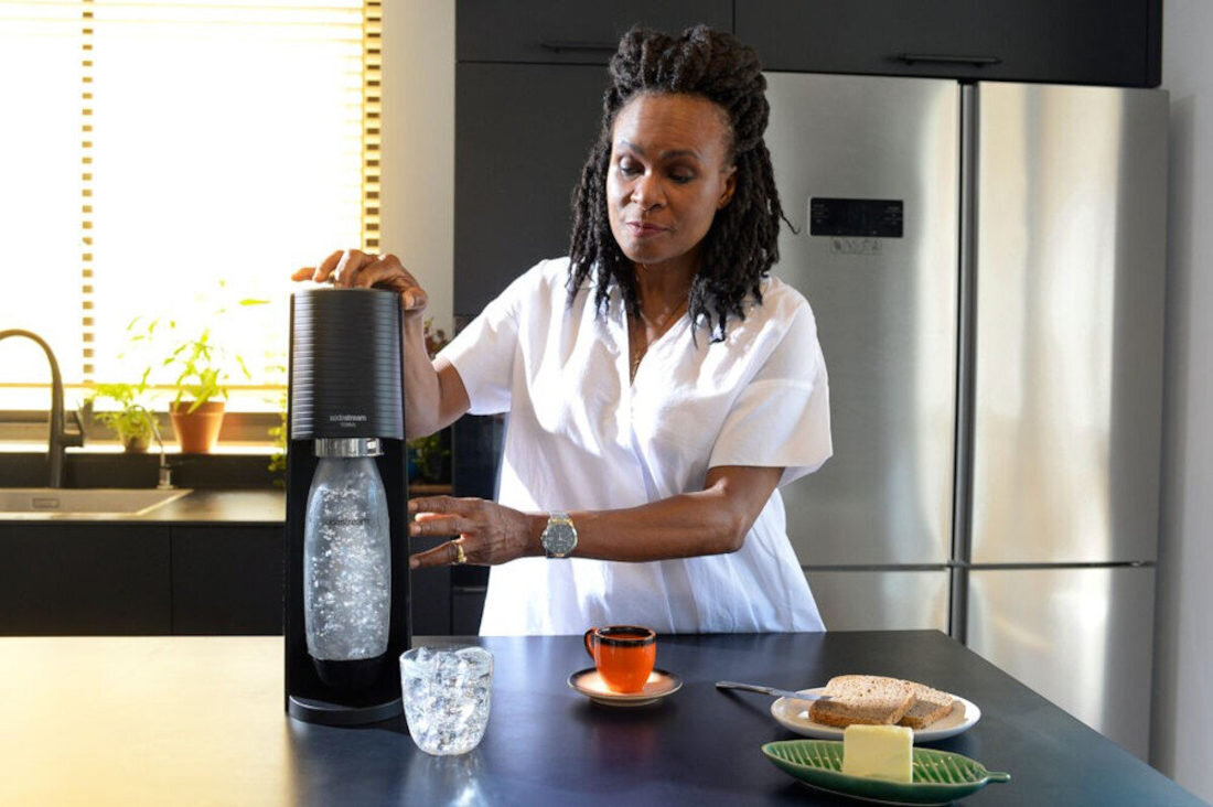 Na blacie kuchennym stoi saturator z butelką wypełnioną wodą gazowaną. Obok znajduje się szklanka z lodem, filiżanka z kawą oraz talerz z pieczywem i masłem.