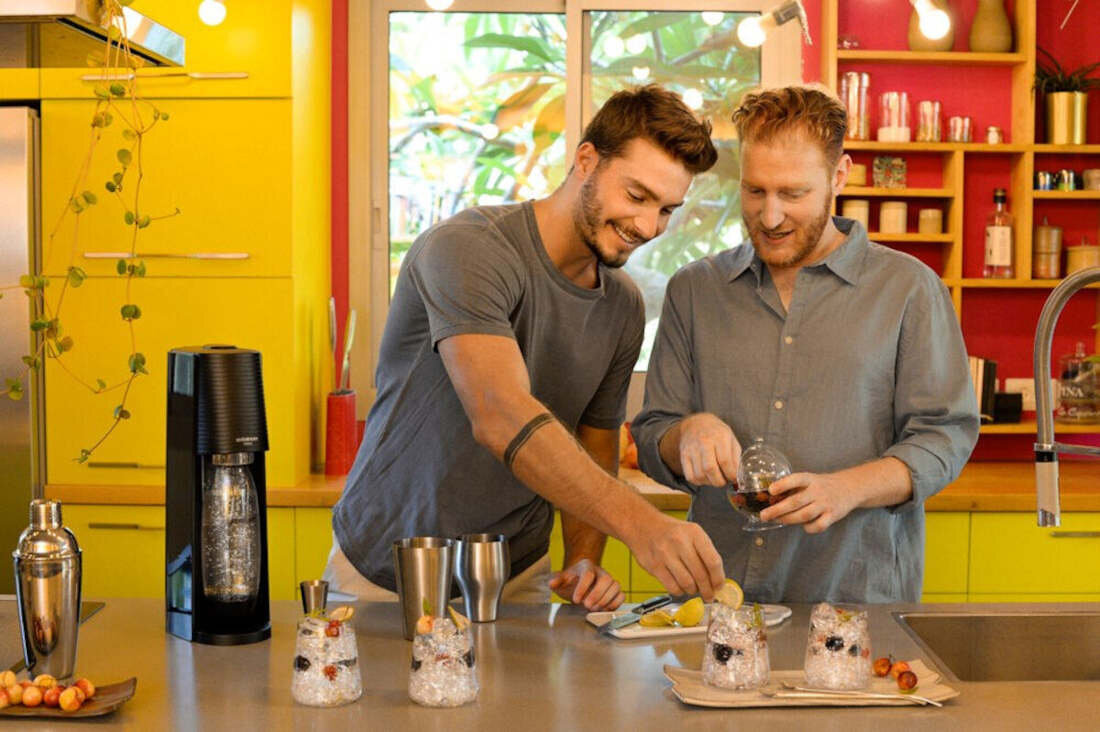 Na blacie kuchennym stoi czarny saturator oraz kilka szklanek z lodem i owocami. W tle widać żółte szafki i okno z widokiem na rośliny.