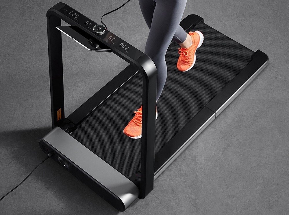 Osoba biegnie na bieżni elektrycznej z czarną powierzchnią do biegania i wyświetlaczem pokazującym dane. Osoba ma na sobie pomarańczowe buty sportowe.