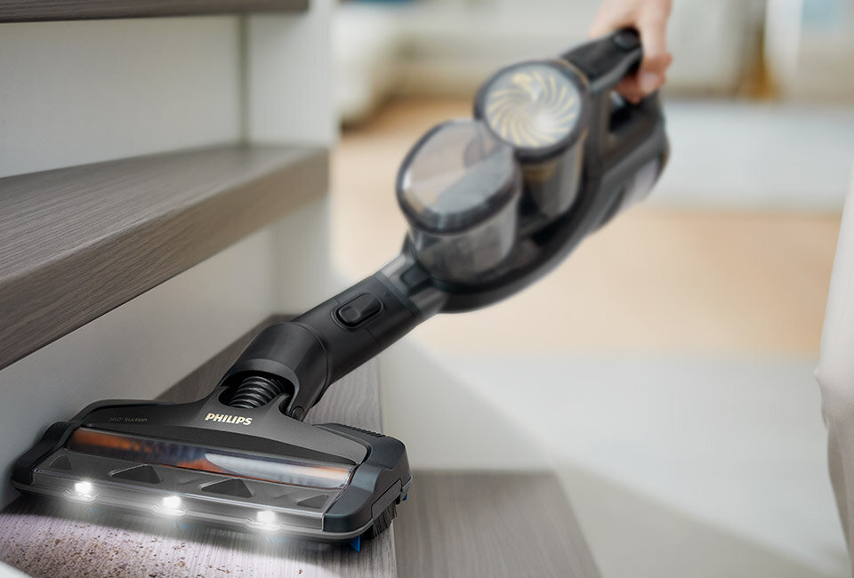 Odkurzacz ręczny z logo PHILIPS, używany do czyszczenia schodów. Urządzenie posiada przezroczysty pojemnik na kurz i jest wyposażone w oświetlenie LED na szczotce.