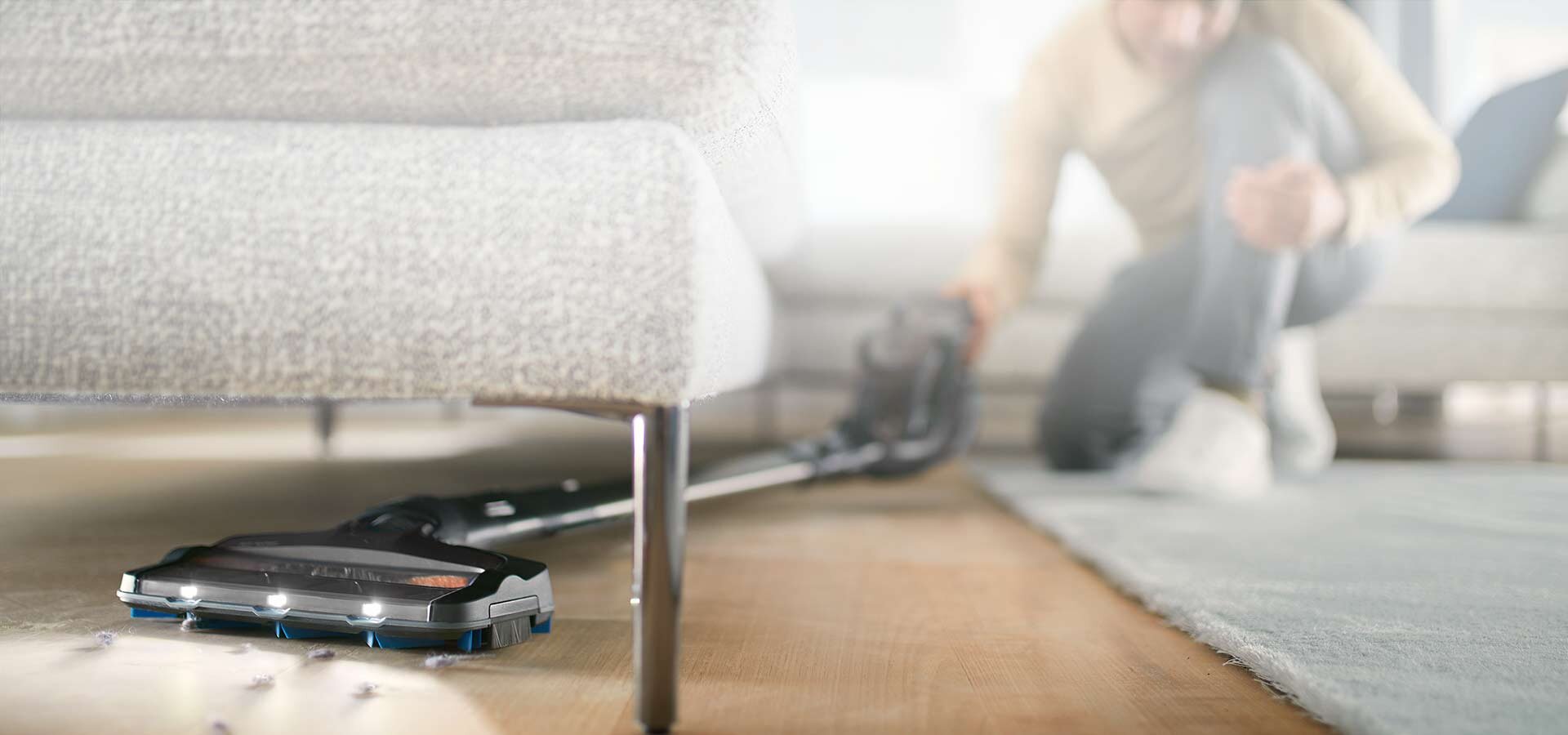 Odkurzacz z funkcją mopowania znajduje się pod sofą, a osoba obsługuje go z boku. Na podłodze widoczne są drobne zanieczyszczenia, które odkurzacz zbiera.