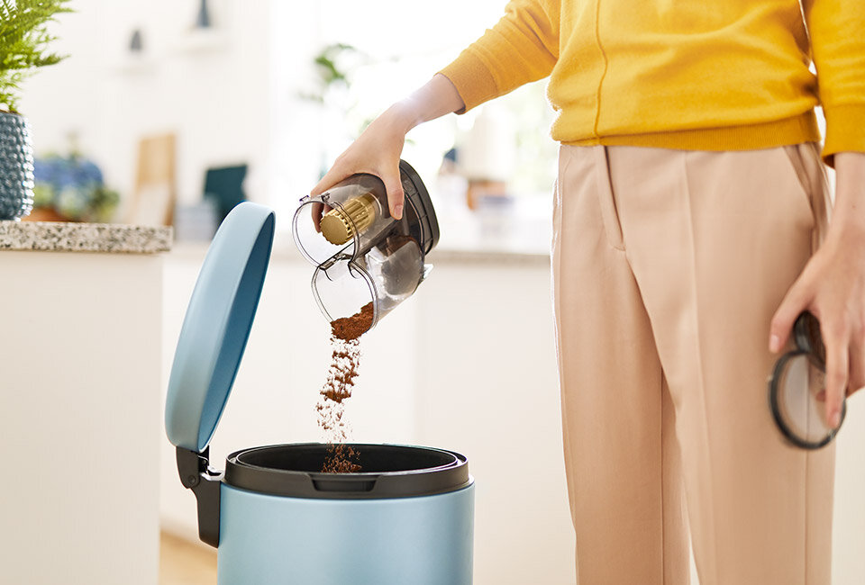Osoba opróżnia pojemnik z odkurzacza do kosza na śmieci. Kosz ma niebieski kolor i otwartą pokrywę.