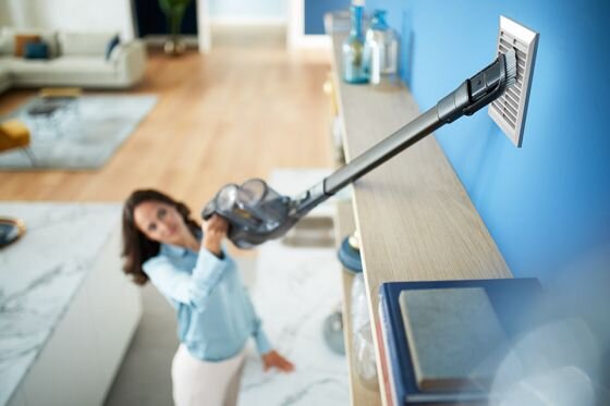 Osoba używa odkurzacza pionowego do czyszczenia kratki wentylacyjnej na ścianie. W tle widać nowoczesne wnętrze z jasną podłogą i meblami.