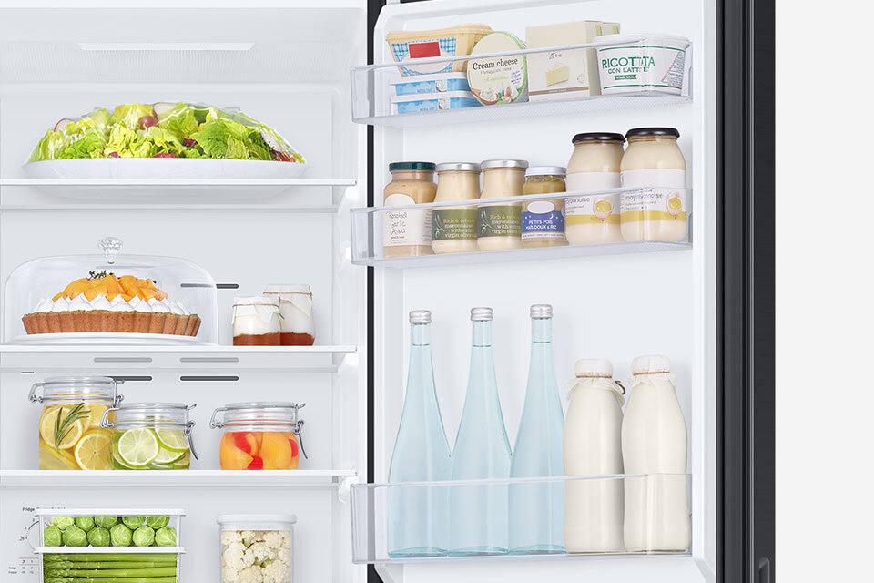 Wnętrze lodówki z widocznymi półkami wypełnionymi różnorodnymi produktami spożywczymi, takimi jak sałata, ciasto, słoiki z owocami, butelki z napojami oraz opakowania z nabiałem. Na drzwiach lodówki znajdują się pojemniki z jogurtami i serkami, a także butelki z mlekiem i wodą.