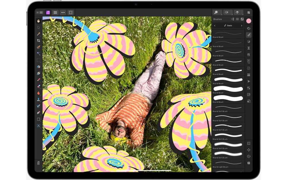 Tablet wyświetla aplikację graficzną z kolorowymi, abstrakcyjnymi kwiatami na tle zdjęcia osoby leżącej na trawie. Po prawej stronie ekranu widoczny jest panel narzędziowy z różnymi opcjami pędzli.