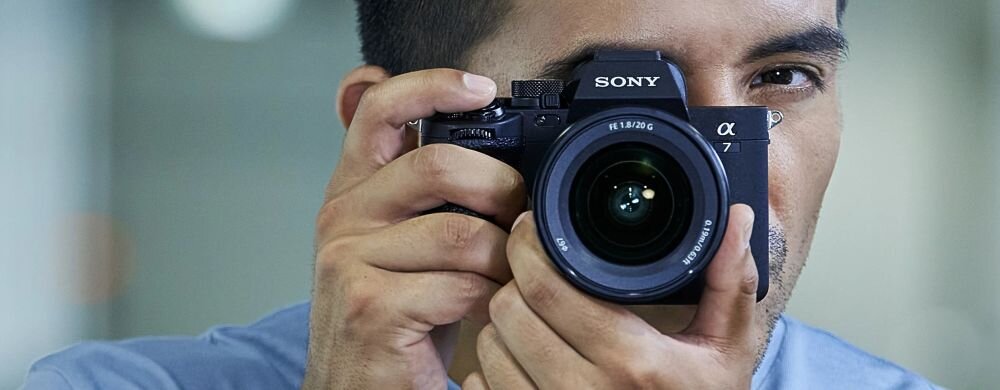 Osoba trzyma czarny aparat bezlusterkowy marki SONY z widocznym obiektywem i logo \'SONY\' na górze. Aparat ma oznaczenie \'α7\'.