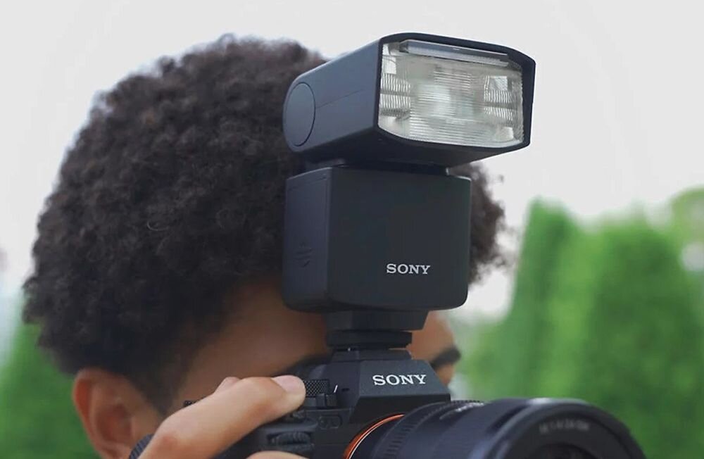Osoba trzyma aparat fotograficzny z zamontowaną lampą błyskową, na której widoczne jest logo \'SONY\'. W tle znajdują się rozmyte zielone elementy, prawdopodobnie roślinność.