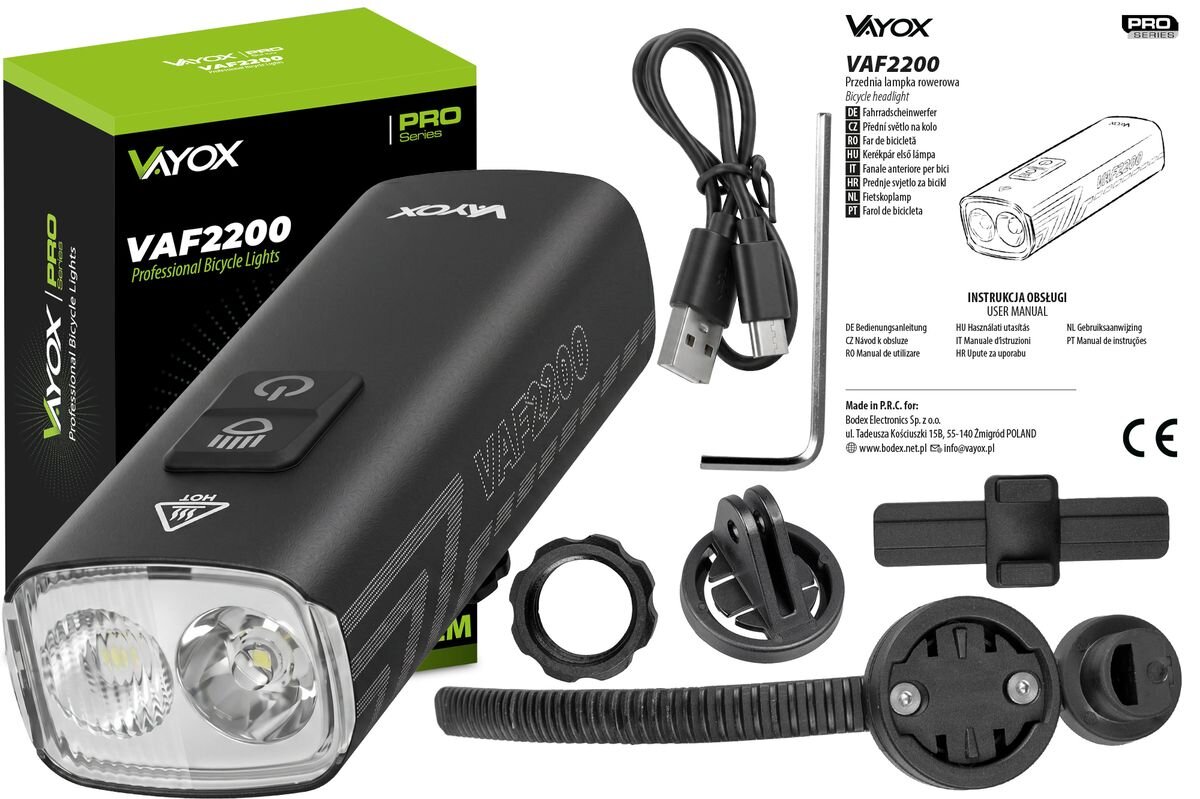 Na obrazie znajduje się lampka rowerowa VAYOX VAF2200 wraz z akcesoriami, takimi jak kabel USB, klucz imbusowy i elementy montażowe. Widoczne są również opakowanie produktu oraz instrukcja obsługi z tekstem w kilku językach.