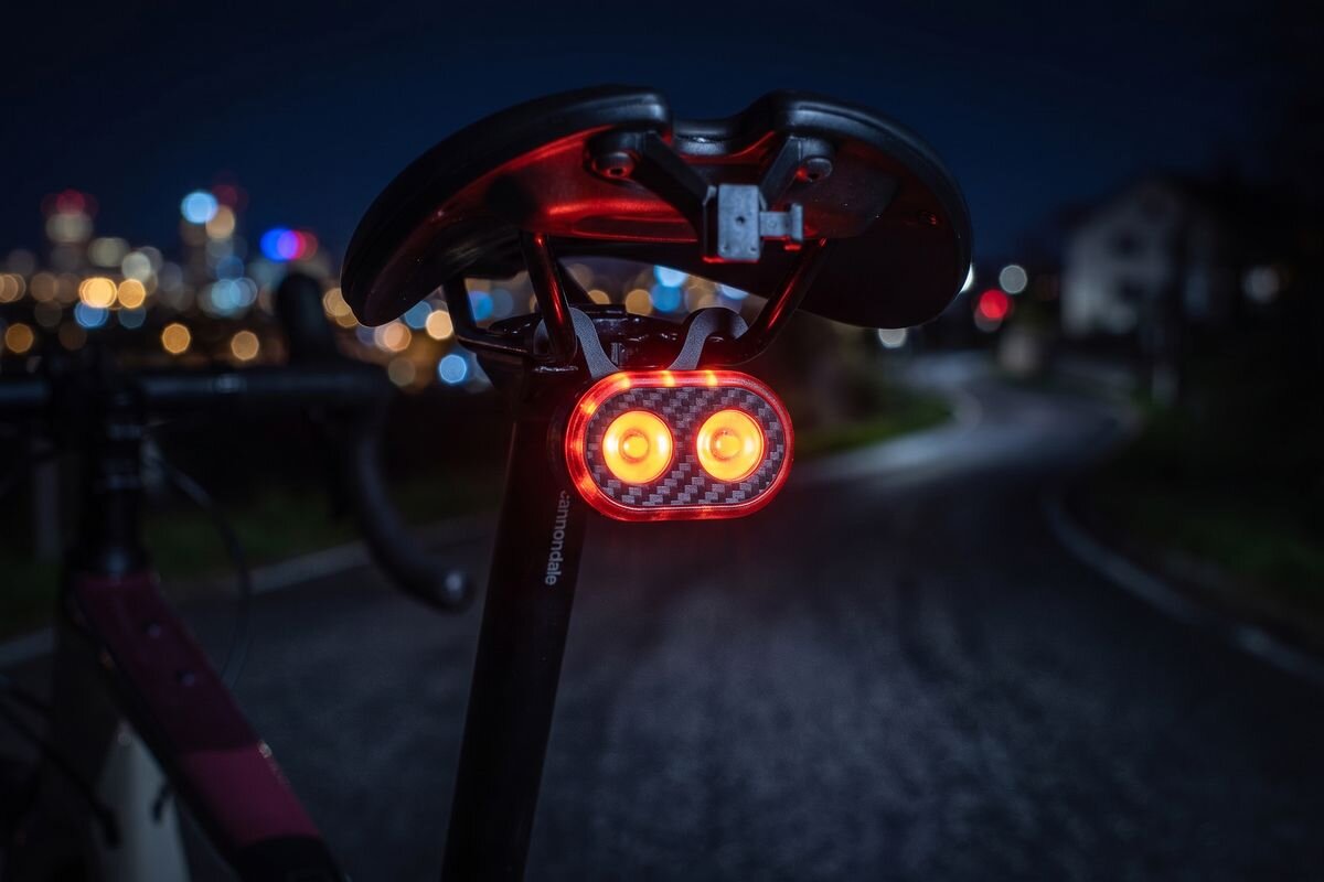 Na zdjęciu widać tylną lampkę rowerową emitującą czerwone światło, zamontowaną pod siodełkiem roweru. W tle rozmyte światła miasta nocą.