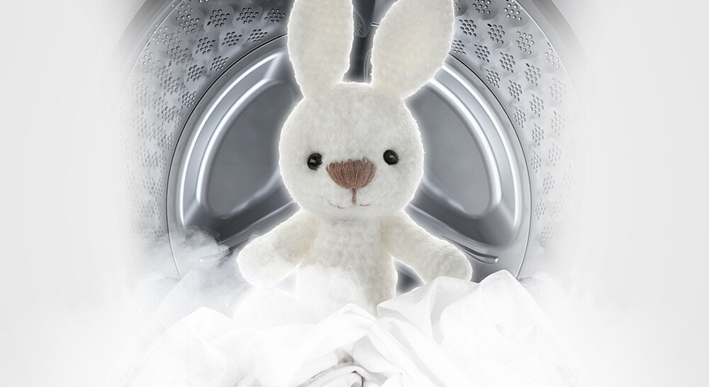 Pluszowy królik znajduje się wewnątrz bębna pralki, otoczony białą parą i tkaninami. Wnętrze bębna jest metaliczne z charakterystycznymi otworami.